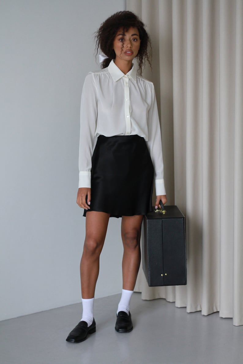 May include: A woman wearing a white button-down shirt with a black skirt and black loafers. She is holding a black briefcase. The outfit is simple and elegant.