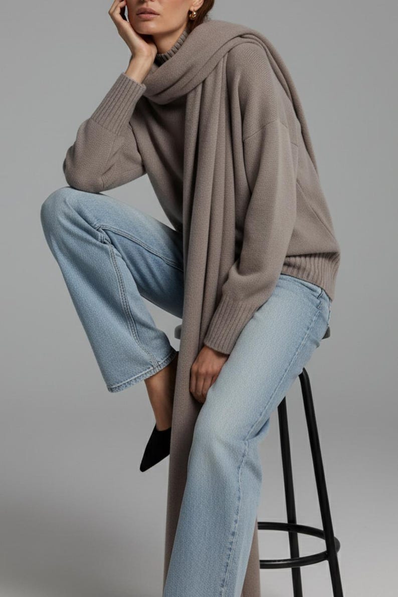 May include: A woman's outfit featuring a taupe-colored sweater and scarf paired with light blue jeans. The outfit is completed with black shoes. The woman is sitting on a black stool.