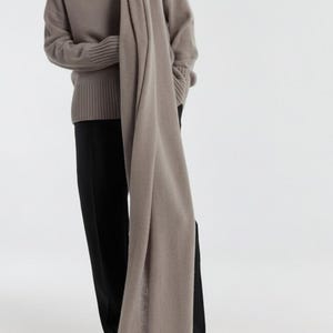 May include: A woman wearing a taupe-colored sweater and scarf, paired with black pants and shoes. The scarf is draped around the neck and over the shoulders, creating a layered look. The outfit is simple and elegant.