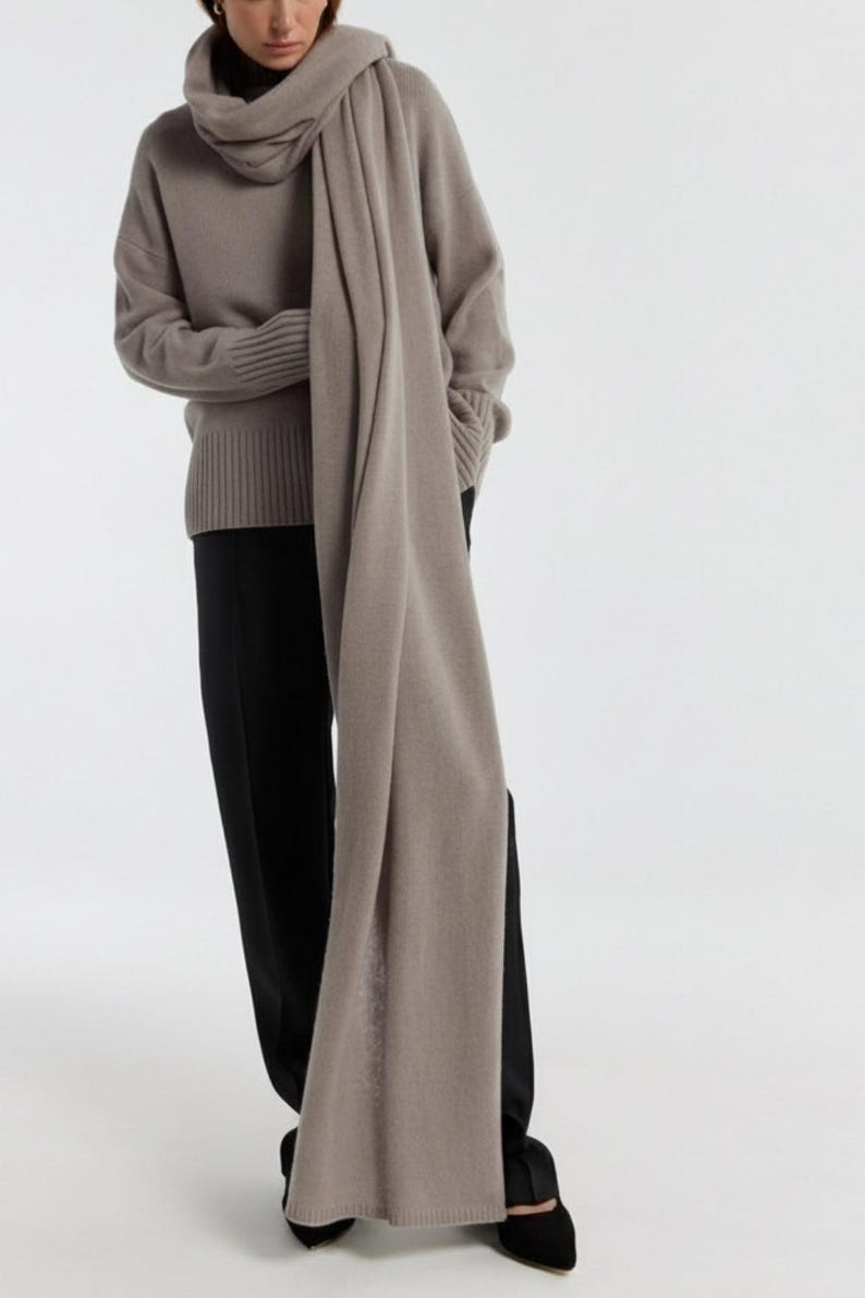 May include: A woman wearing a taupe-colored sweater and scarf, paired with black pants and shoes. The scarf is draped around the neck and over the shoulders, creating a layered look. The outfit is simple and elegant.