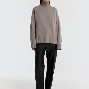 May include: A woman wearing a taupe turtleneck sweater and black trousers. The sweater has a ribbed hem and cuffs. The trousers are straight-legged. The woman is also wearing black pointed-toe shoes.
