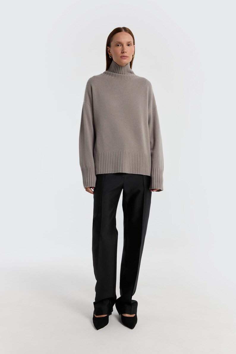 May include: A woman wearing a taupe turtleneck sweater and black trousers. The sweater has a ribbed hem and cuffs. The trousers are straight-legged. The woman is also wearing black pointed-toe shoes.