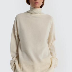 May include: A cream-colored turtleneck sweater with long sleeves. The sweater has a ribbed hem and cuffs. The model is wearing cream-colored pants. The background is a solid white.