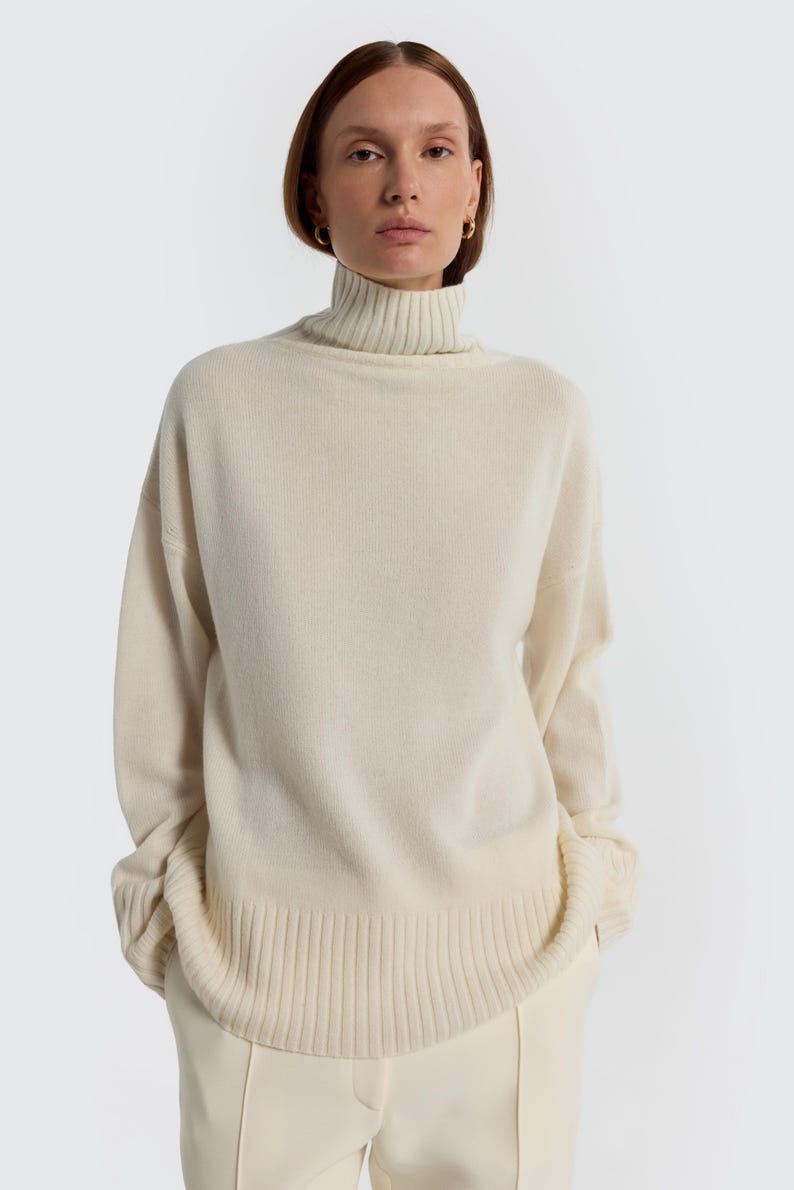 May include: A cream-colored turtleneck sweater with long sleeves. The sweater has a ribbed hem and cuffs. The model is wearing cream-colored pants. The background is a solid white.