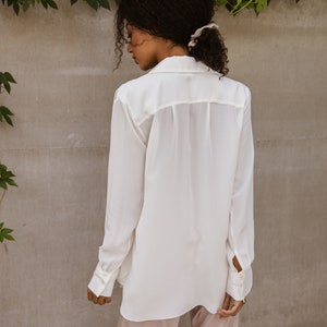 May include: A white button-down shirt with long sleeves and a relaxed fit. The shirt is paired with light pink satin pants. The model is wearing a light-colored scrunchie in their hair. The background is a neutral gray wall with green vines.