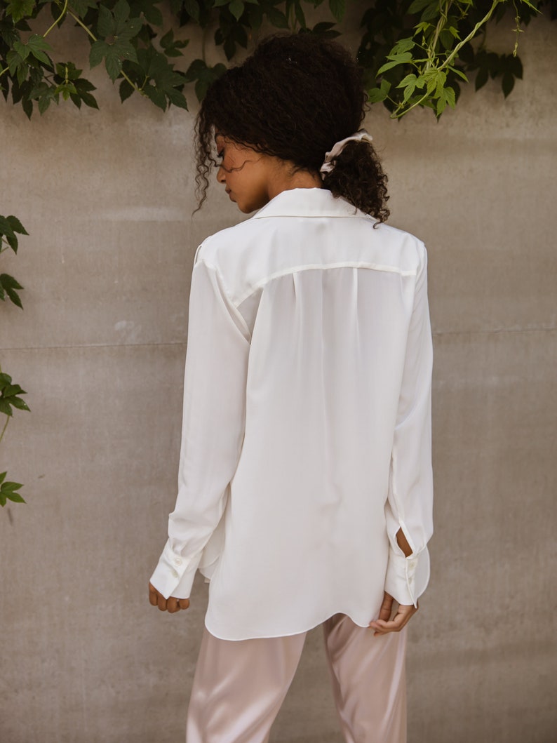 May include: A white button-down shirt with long sleeves and a relaxed fit. The shirt is paired with light pink satin pants. The model is wearing a light-colored scrunchie in their hair. The background is a neutral gray wall with green vines.