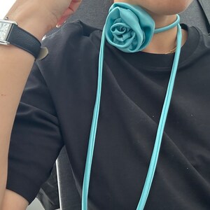 May include: A light blue satin rose choker necklace with a long, thin strap. The necklace is worn by a person with a black shirt.