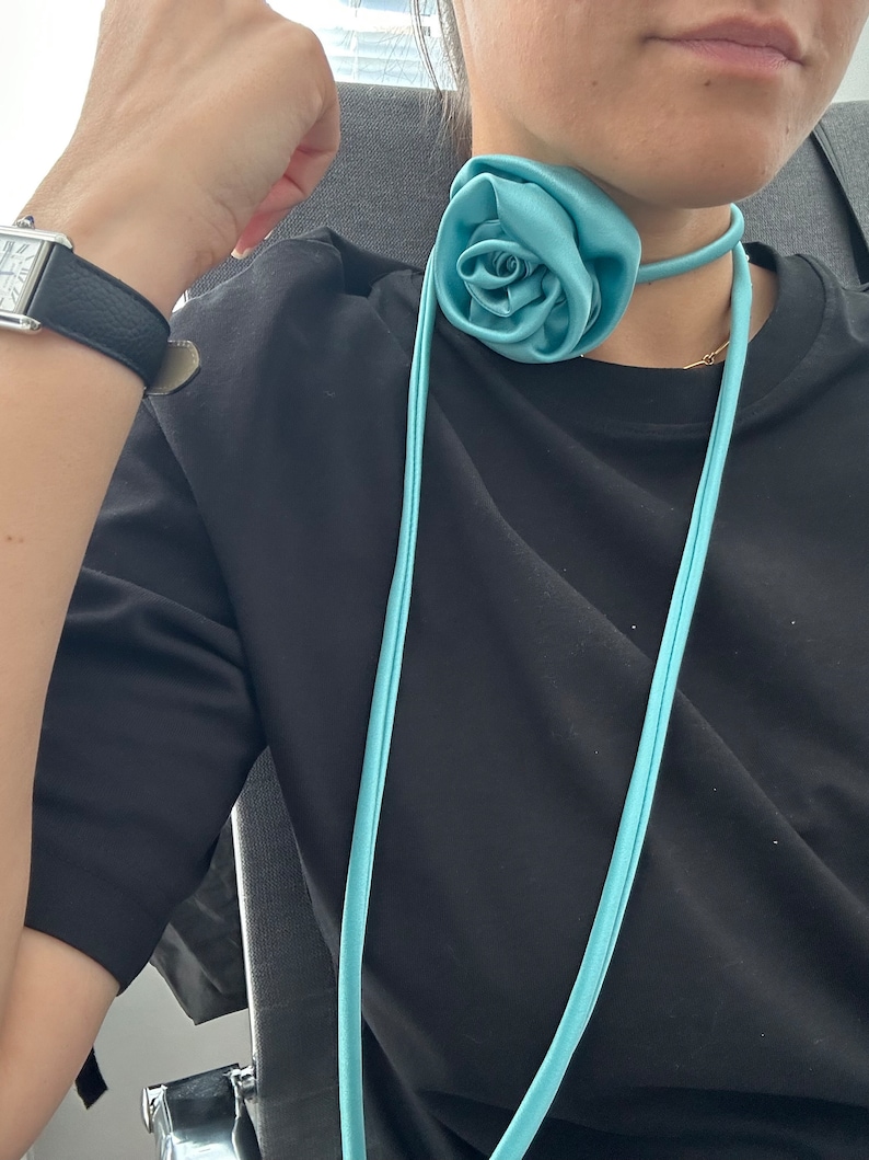 May include: A light blue satin rose choker necklace with a long, thin strap. The necklace is worn by a person with a black shirt.