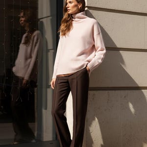May include: A woman wearing a light pink turtleneck sweater and dark brown trousers. The sweater has long sleeves and a ribbed hem. The trousers are wide-legged. The woman is standing next to a building with a glass window.