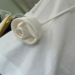 May include: A white satin rose with a long stem, attached to a white t-shirt.