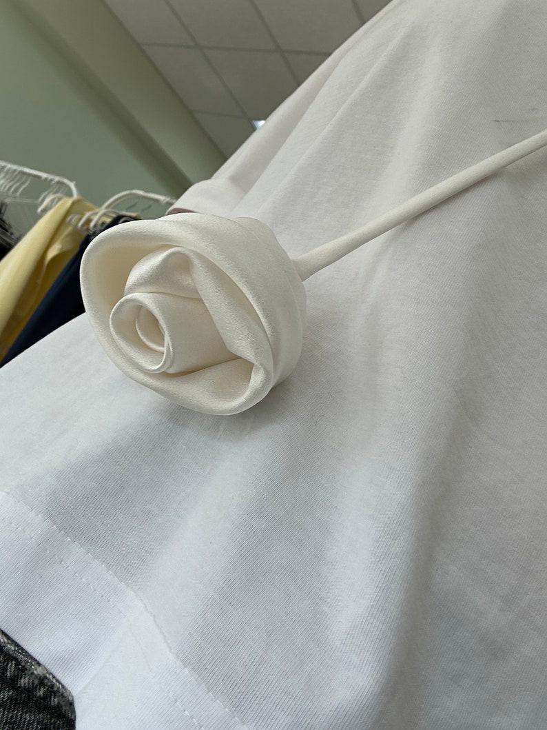 May include: A white satin rose with a long stem, attached to a white t-shirt.