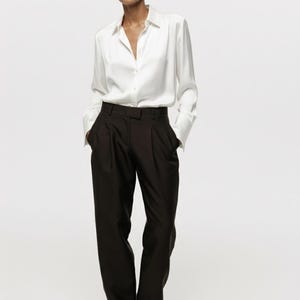 Puede incluir: Una camisa blanca de manga larga con cuello cl&aacute;sico y pu&ntilde;os, combinada con pantalones de color marr&oacute;n oscuro de talle alto con frente plisado, perneras rectas y un corte entallado y limpio con cierre de cremallera.