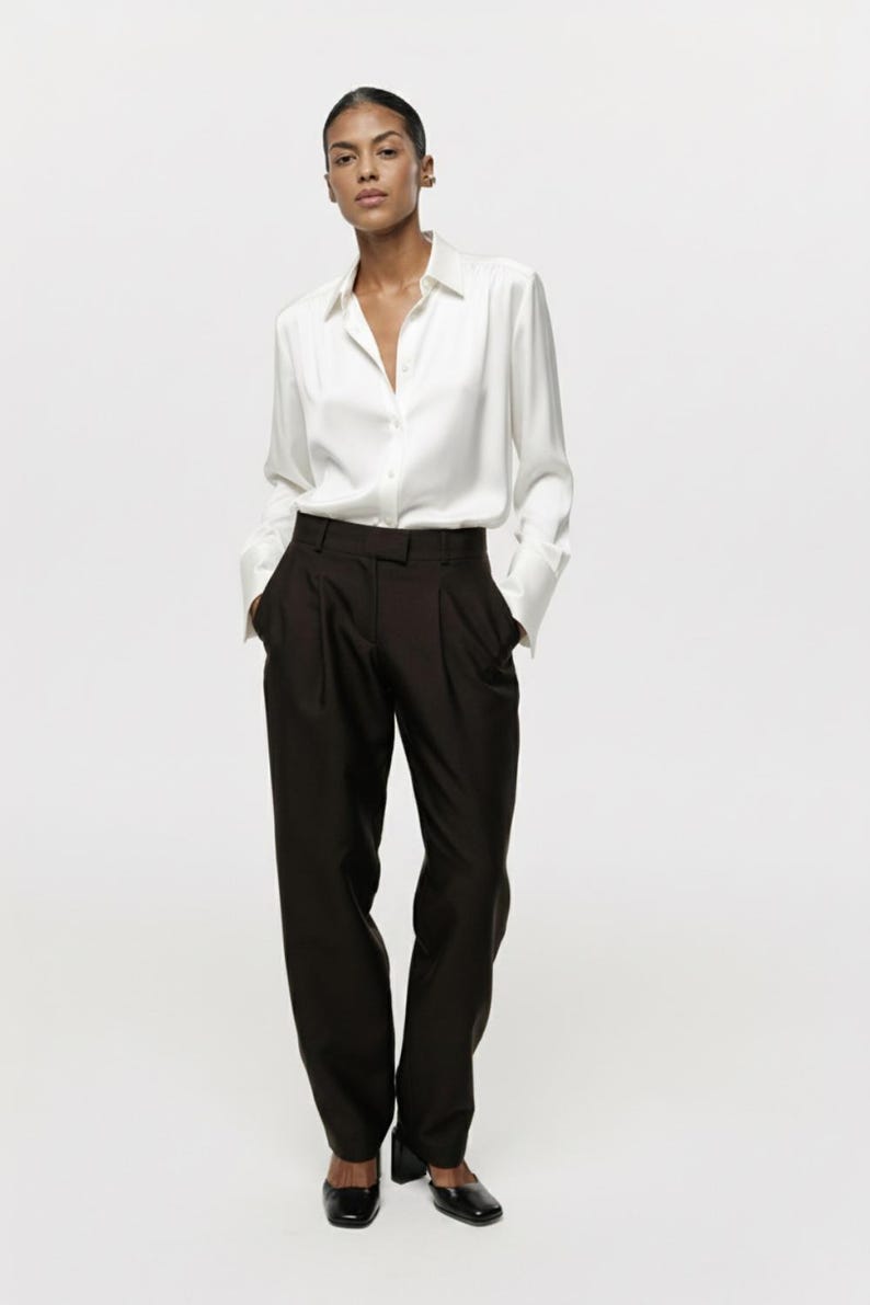 Puede incluir: Una camisa blanca de manga larga con cuello cl&aacute;sico y pu&ntilde;os, combinada con pantalones de color marr&oacute;n oscuro de talle alto con frente plisado, perneras rectas y un corte entallado y limpio con cierre de cremallera.