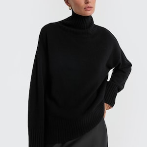 May include: A black turtleneck sweater with long sleeves. The sweater has a ribbed hem and cuffs. The model is wearing a black skirt. The background is plain white. This is a classic, versatile piece of clothing.