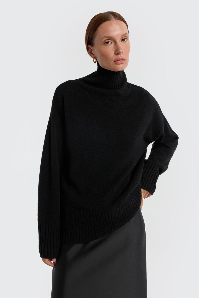 May include: A black turtleneck sweater with long sleeves. The sweater has a ribbed hem and cuffs. The model is wearing a black skirt. The background is plain white. This is a classic, versatile piece of clothing.