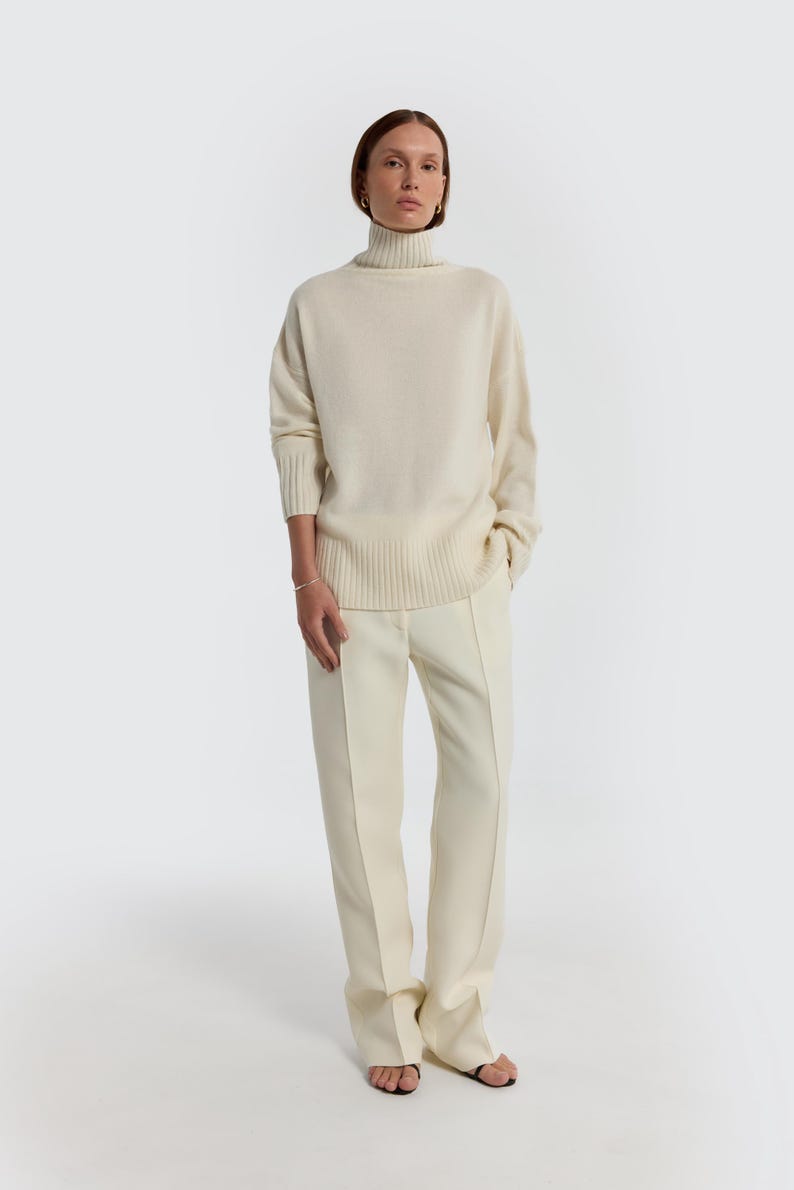 May include: A cream-colored turtleneck sweater and matching pants. The sweater has a ribbed hem and cuffs. The pants are a straight-leg style. The outfit is a neutral, minimalist ensemble.