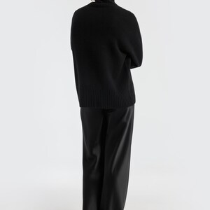 May include: A person wearing a black turtleneck sweater and black satin pants. The sweater has a ribbed hem and cuffs. The pants have a wide leg design. The background is plain white.