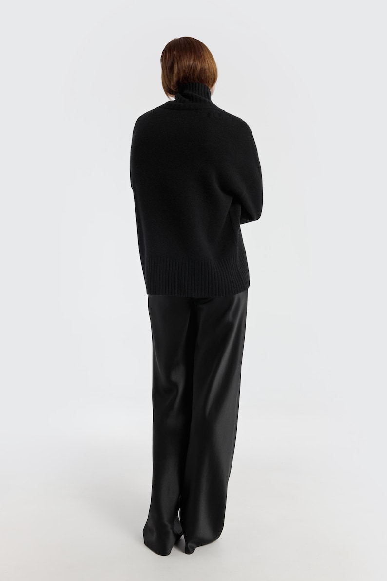 May include: A person wearing a black turtleneck sweater and black satin pants. The sweater has a ribbed hem and cuffs. The pants have a wide leg design. The background is plain white.