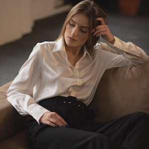 Puede incluir: Una camisa blanca de manga larga con cuello cl&aacute;sico y pu&ntilde;os, combinada con pantalones negros de talle alto que incluyen un cintur&oacute;n ancho de tela con hebilla rectangular. La camisa presenta un acabado liso y ligeramente brillante.