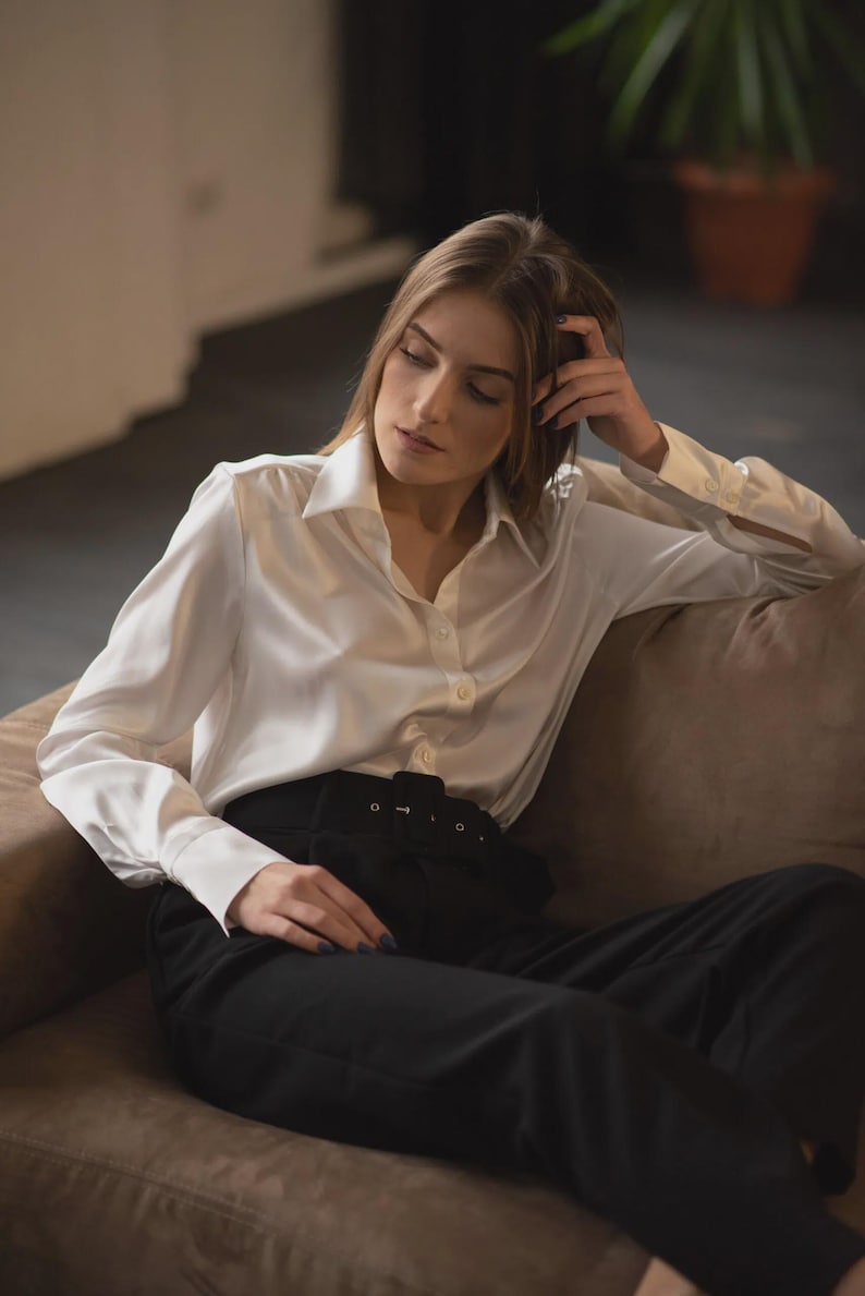 Puede incluir: Una camisa blanca de manga larga con cuello cl&aacute;sico y pu&ntilde;os, combinada con pantalones negros de talle alto que incluyen un cintur&oacute;n ancho de tela con hebilla rectangular. La camisa presenta un acabado liso y ligeramente brillante.