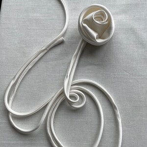 May include: A white satin ribbon tied into a rose shape with the ends of the ribbon forming a loose circle.