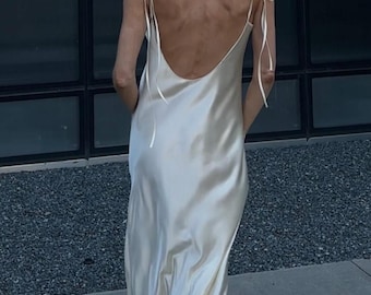 May include: A cream-coloured, floor-length slip dress with a low back and delicate tie straps. The dress is made of a silky material that drapes elegantly. The model is standing on a concrete surface.