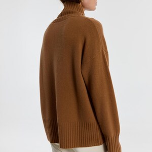 May include: A brown turtleneck sweater with long sleeves. The sweater has a ribbed hem and cuffs. The model is wearing the sweater with white pants. The sweater is made of a soft, warm material.