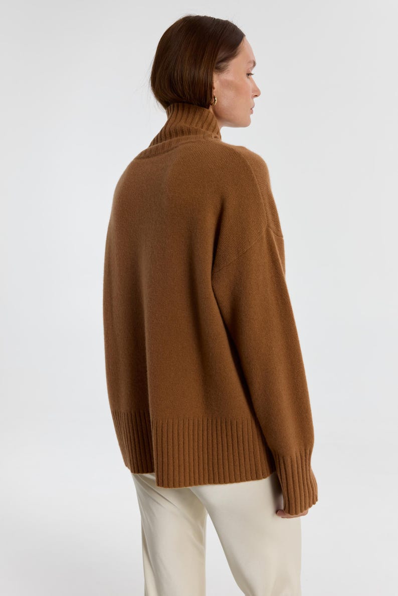 May include: A brown turtleneck sweater with long sleeves. The sweater has a ribbed hem and cuffs. The model is wearing the sweater with white pants. The sweater is made of a soft, warm material.