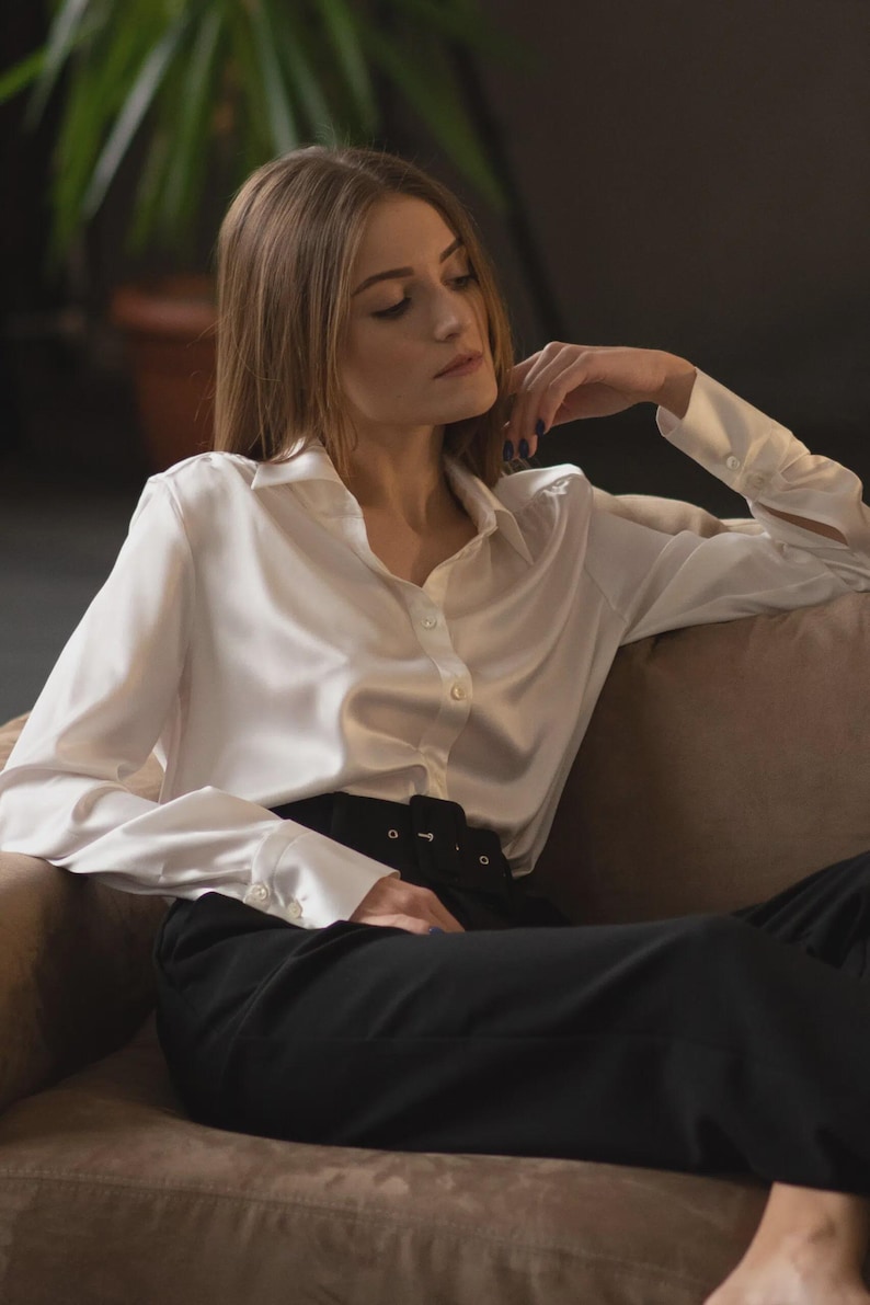 Puede incluir: Una camisa blanca de manga larga con cuello cl&aacute;sico y pu&ntilde;os, combinada con pantalones negros de talle alto que incluyen un cintur&oacute;n ancho con hebilla rectangular. La camisa tiene un acabado satinado suave y una tapeta frontal con botones.