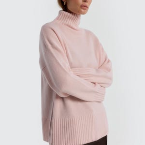 May include: A light pink turtleneck sweater. The sweater has a ribbed hem and cuffs, and a relaxed fit. The model is wearing a dark brown skirt. The background is plain white.