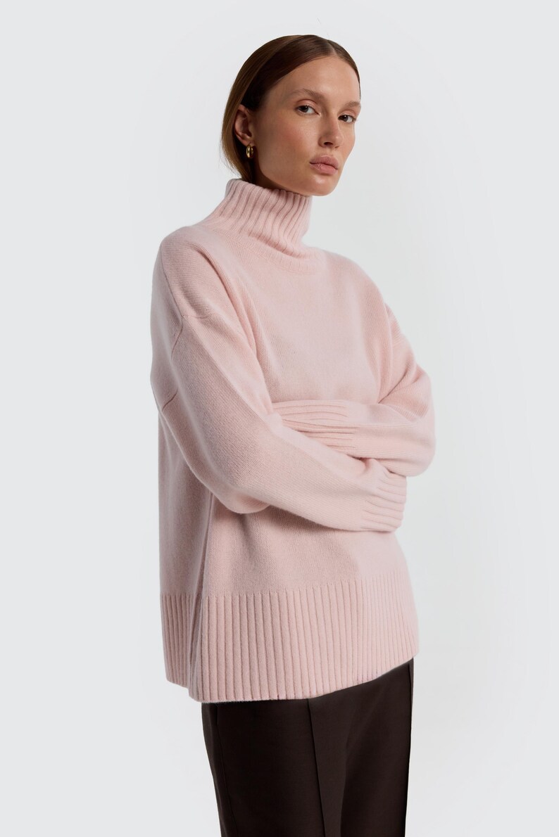 May include: A light pink turtleneck sweater. The sweater has a ribbed hem and cuffs, and a relaxed fit. The model is wearing a dark brown skirt. The background is plain white.