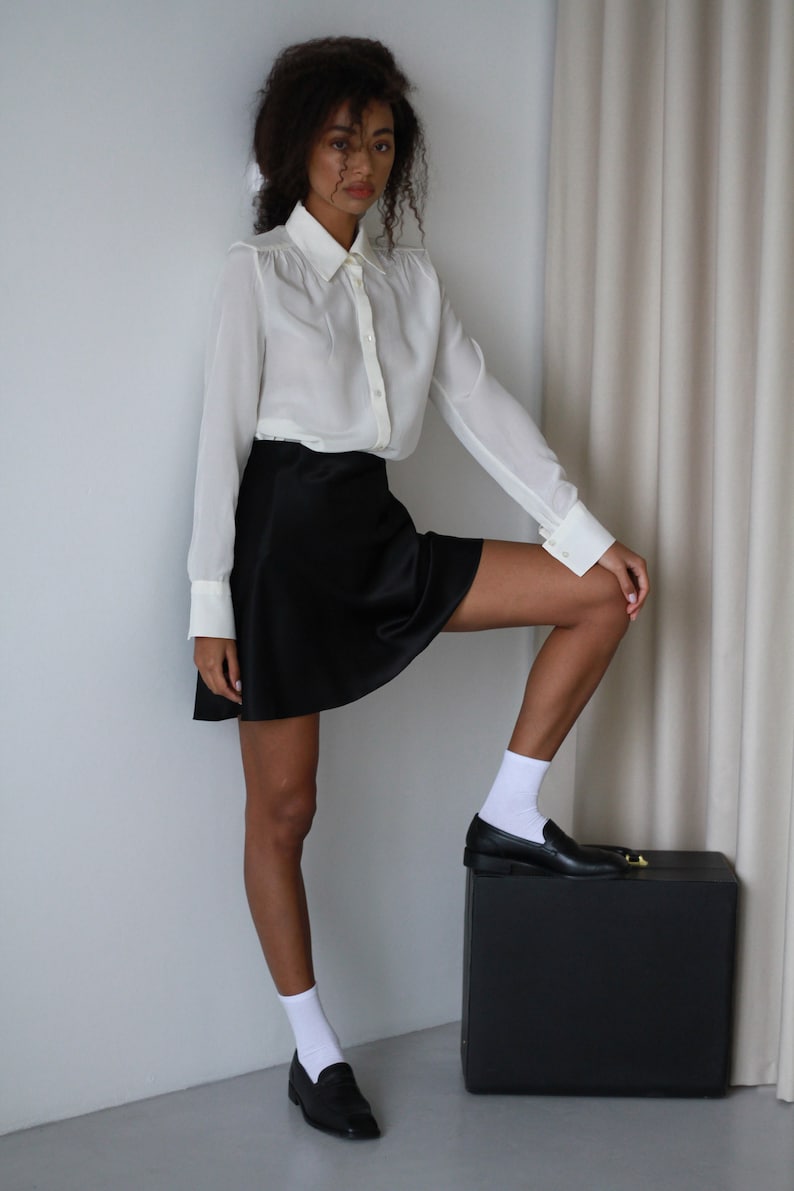 May include: A woman wearing a white button-down shirt with a black mini skirt, white socks, and black loafers. The woman is standing with one leg up on a black box. The shirt has long sleeves and a collared neckline.
