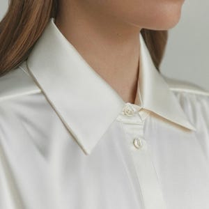 Puede incluir: Una camisa abotonada de color crema con un acabado suave y satinado, que presenta un cuello puntiagudo cl&aacute;sico y una tapeta frontal con botones peque&ntilde;os, redondos y claros. La tela tiene un brillo sutil y una ca&iacute;da fluida.