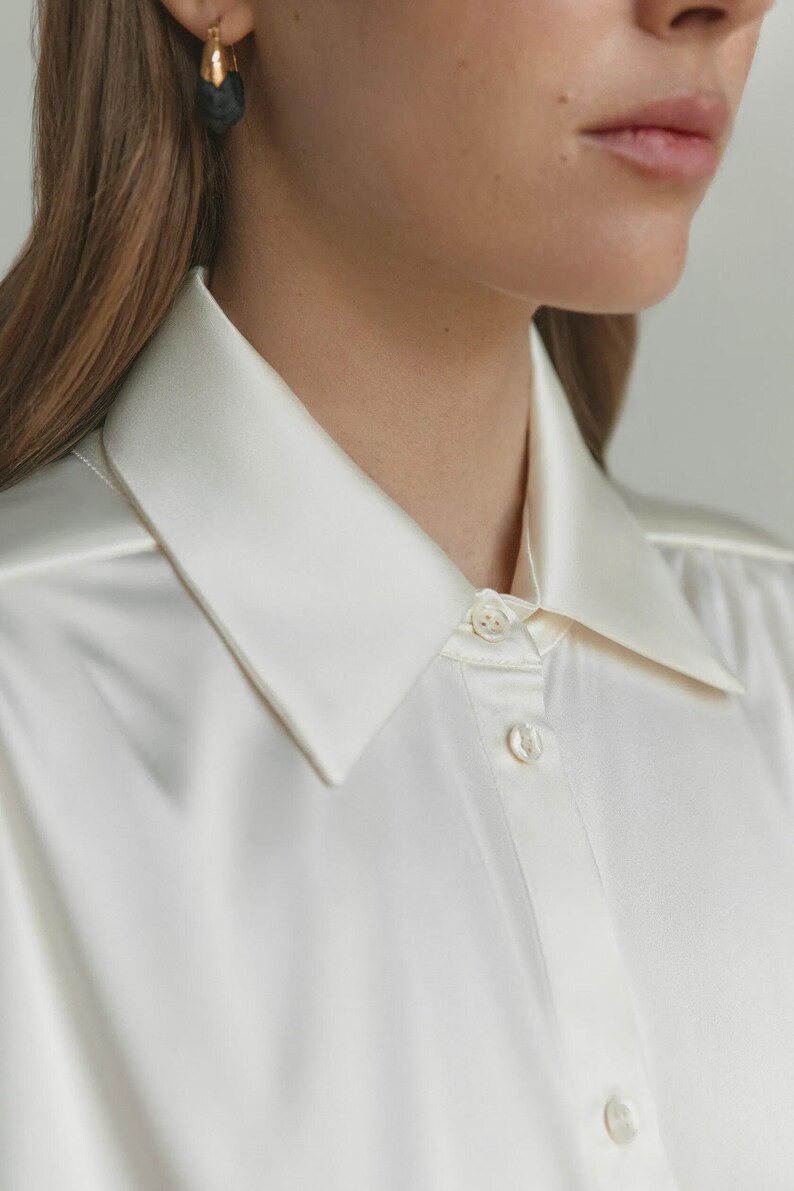 Puede incluir: Una camisa abotonada de color crema con un acabado suave y satinado, que presenta un cuello puntiagudo cl&aacute;sico y una tapeta frontal con botones peque&ntilde;os, redondos y claros. La tela tiene un brillo sutil y una ca&iacute;da fluida.