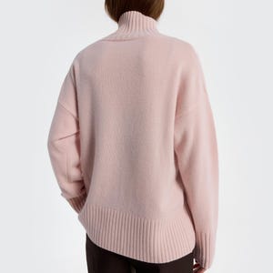 May include: A light pink turtleneck sweater. The sweater has a ribbed hem and cuffs, and a relaxed fit. The model is wearing dark brown pants. The sweater is made of a soft, possibly wool, material.