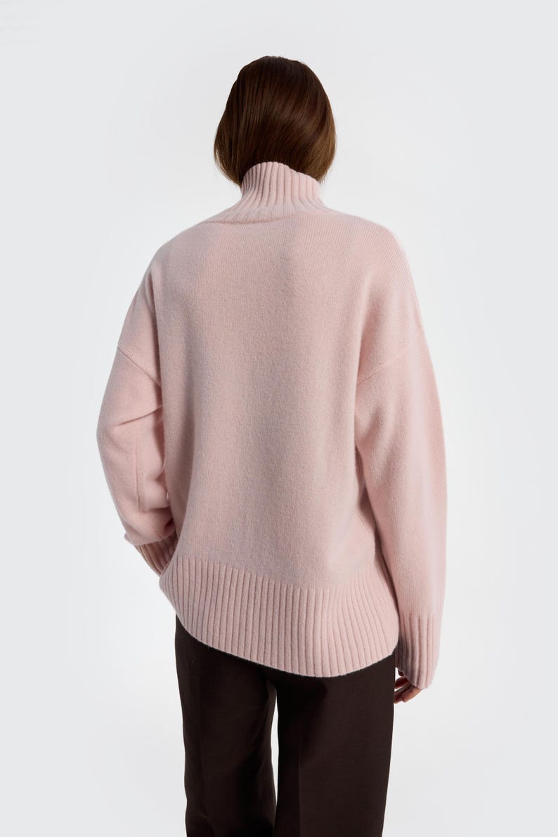 May include: A light pink turtleneck sweater. The sweater has a ribbed hem and cuffs, and a relaxed fit. The model is wearing dark brown pants. The sweater is made of a soft, possibly wool, material.