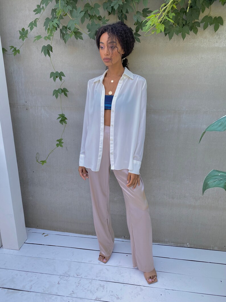 May include: A cream-colored button-down shirt paired with blush-colored wide-leg pants. The outfit includes a blue and white patterned top and nude-colored square-toe sandals. A gold necklace adds a touch of elegance.