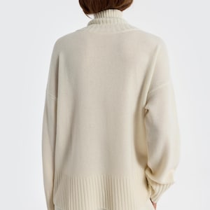 May include: Cream-colored turtleneck sweater with ribbed detailing at the hem and cuffs. The sweater is a neutral tone, offering a versatile wardrobe staple. The model is wearing matching cream-colored pants.