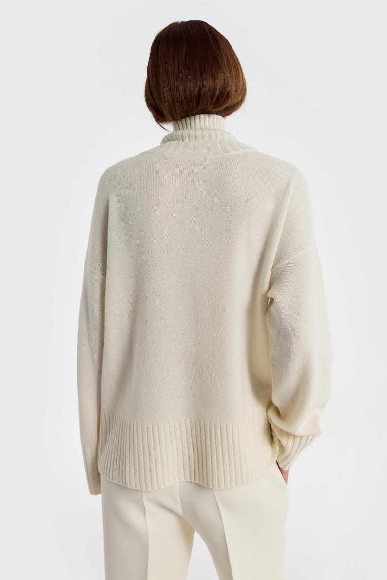 May include: Cream-colored turtleneck sweater with ribbed detailing at the hem and cuffs. The sweater is a neutral tone, offering a versatile wardrobe staple. The model is wearing matching cream-colored pants.