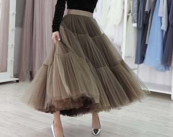 May include: A woman wearing a black long-sleeved top with a square neckline and a brown tulle skirt. She is wearing silver heels and standing in a room with a rack of clothes in the background.