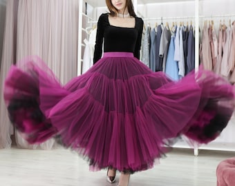 May include: A woman wearing a black velvet top with a square neckline and a long, flowing, layered tulle skirt in a deep magenta color. The skirt is billowing out around her as she stands in a room with a clothing rack in the background.