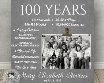 May include: A gray and white 100th birthday sign with a photo of a family. The sign includes the text "100 Years", "1200 months", "36,524 Days", "876,582 Hours", "52,594,920 Minutes", "4 Loving Children", "8 Amazing Grandchildren", "10 Precious Great Grandchildren", "1 Blessed Life", "Splendid Memories", "Happy Birthday", "We Love You", "Mary Elizabeth Stevens", and "April 1, 1922".
