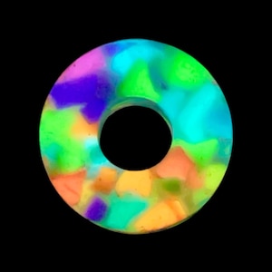 Op de afbeelding: Een cirkelvormig, ringvormig object met een levendig mozaïekpatroon in neonkleuren, waaronder felgroen, oranje, paars en blauw. Het oppervlak vertoont een veelkleurig, gefragmenteerd uiterlijk met een gladde afwerking en een centrale ronde uitsparing.