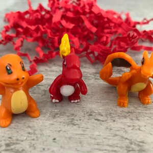 Może przedstawiać: Trzy małe plastikowe figurki postaci Pokemon. Postacie to Charmander, Charmeleon i Charizard. Charmander jest czerwony z ż&oacute;łtym płomieniem na głowie. Charmeleon jest pomarańczowy z ż&oacute;łtym płomieniem na głowie. Charizard jest pomarańczowy z czarnymi skrzydłami i ż&oacute;łtym płomieniem na głowie.