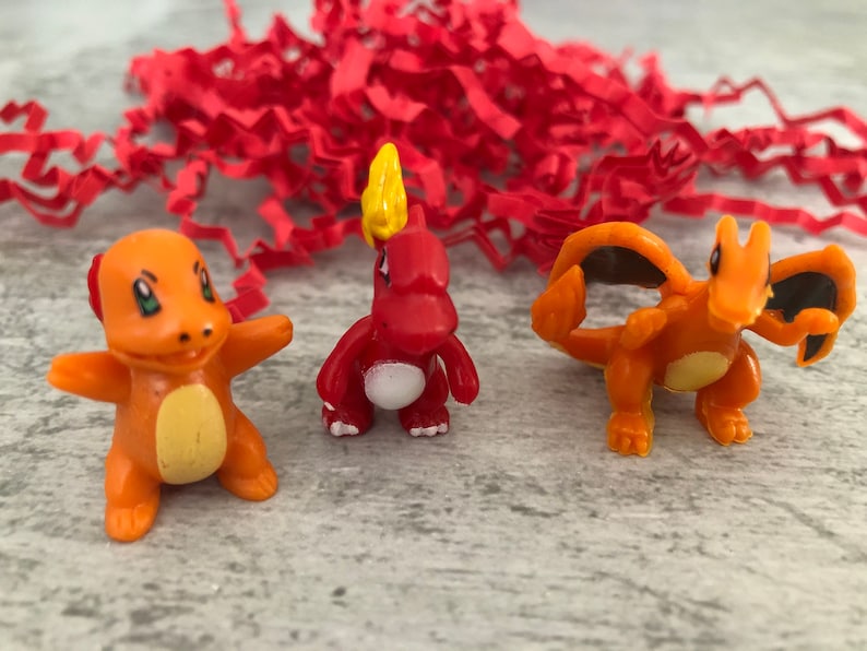Może przedstawiać: Trzy małe plastikowe figurki postaci Pokemon. Postacie to Charmander, Charmeleon i Charizard. Charmander jest czerwony z ż&oacute;łtym płomieniem na głowie. Charmeleon jest pomarańczowy z ż&oacute;łtym płomieniem na głowie. Charizard jest pomarańczowy z czarnymi skrzydłami i ż&oacute;łtym płomieniem na głowie.