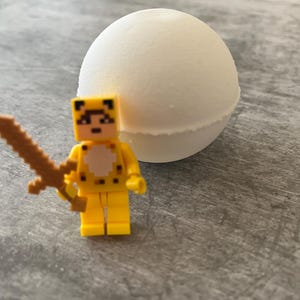 May include: A yellow toy figure with a cheetah-like design and a brown sword stands next to a large, white, spherical bath bomb. The figure has black pixelated details and is made of plastic. The bath bomb has a textured surface.