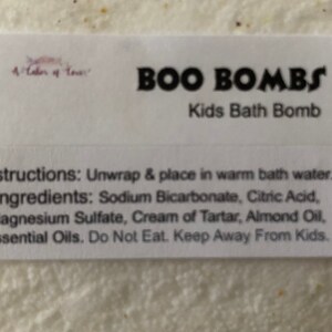Może przedstawiać: Biała etykieta z napisem "BOO BOMBS Kids Bath Bomb". Instrukcje i składniki są wymienione pod tytułem. Składniki obejmują wodorowęglan sodu, kwas cytrynowy i olejki eteryczne.