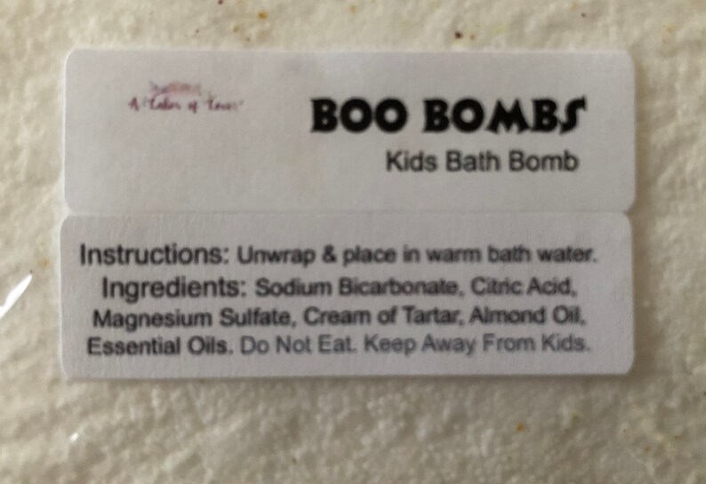 Może przedstawiać: Biała etykieta z napisem "BOO BOMBS Kids Bath Bomb". Instrukcje i składniki są wymienione pod tytułem. Składniki obejmują wodorowęglan sodu, kwas cytrynowy i olejki eteryczne.