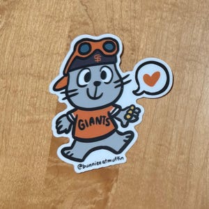 May include: A gray cartoon cat wearing a baseball cap with goggles, an orange jersey with the word "GIANTS" on it, and a heart-shaped speech bubble. The cat is walking and holding a small yellow object in its hand. The caption at the bottom reads "@bunnieeatmuffin".