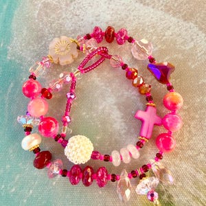 May include: A multi-strand beaded bracelet featuring a variety of pink, clear, and white beads. The design includes a central pink cross charm, a white textured spherical bead, a translucent flower-shaped bead, and faceted crystal accents.